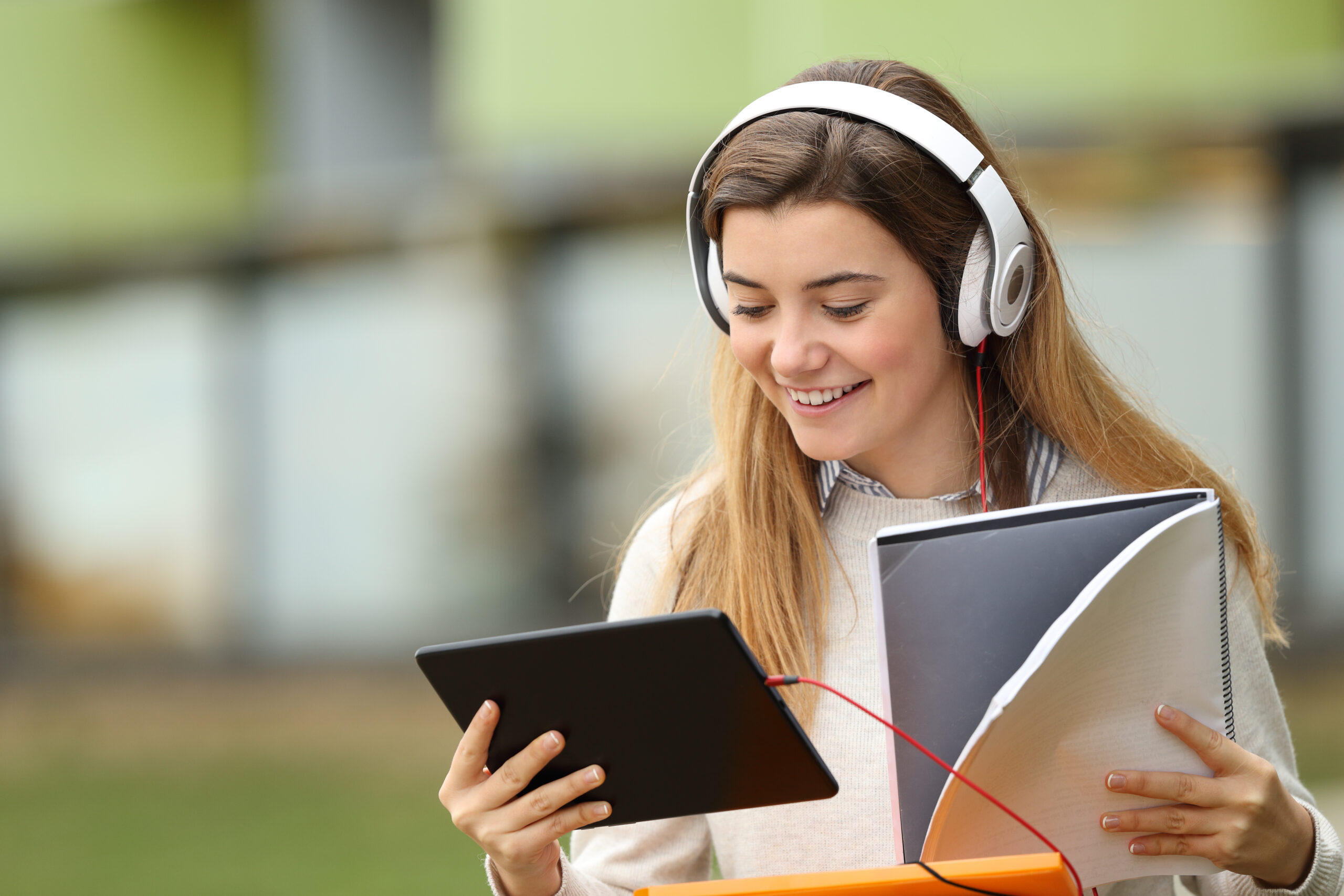 A young woman wearing a white sweater sits in the quad viewing a tablet while wearing wired headphones and holding a notebook in the other hand