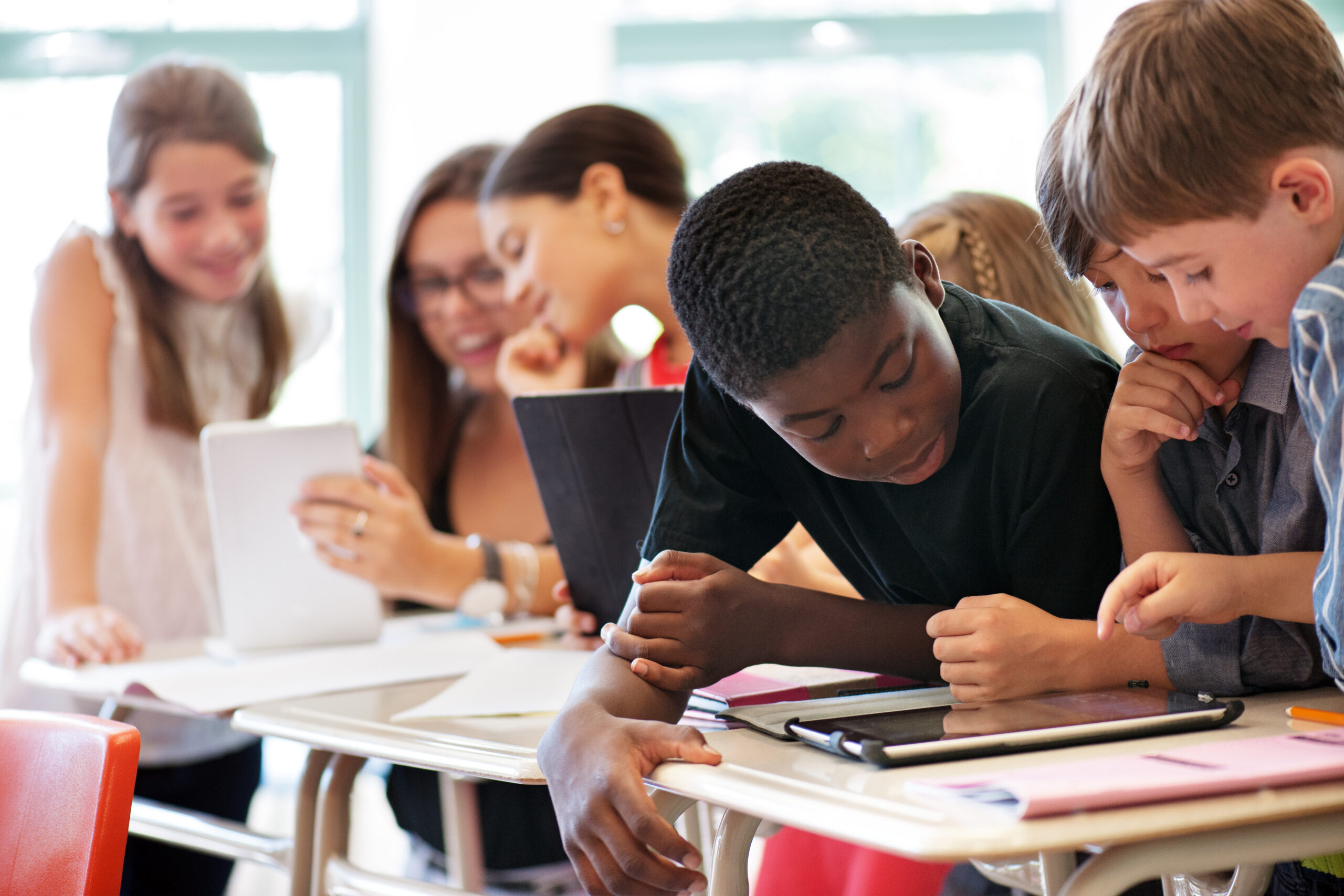 A group of three boys gather around reading a tablet at their desks. In the background a teacher assists two girls with reading a tablet.