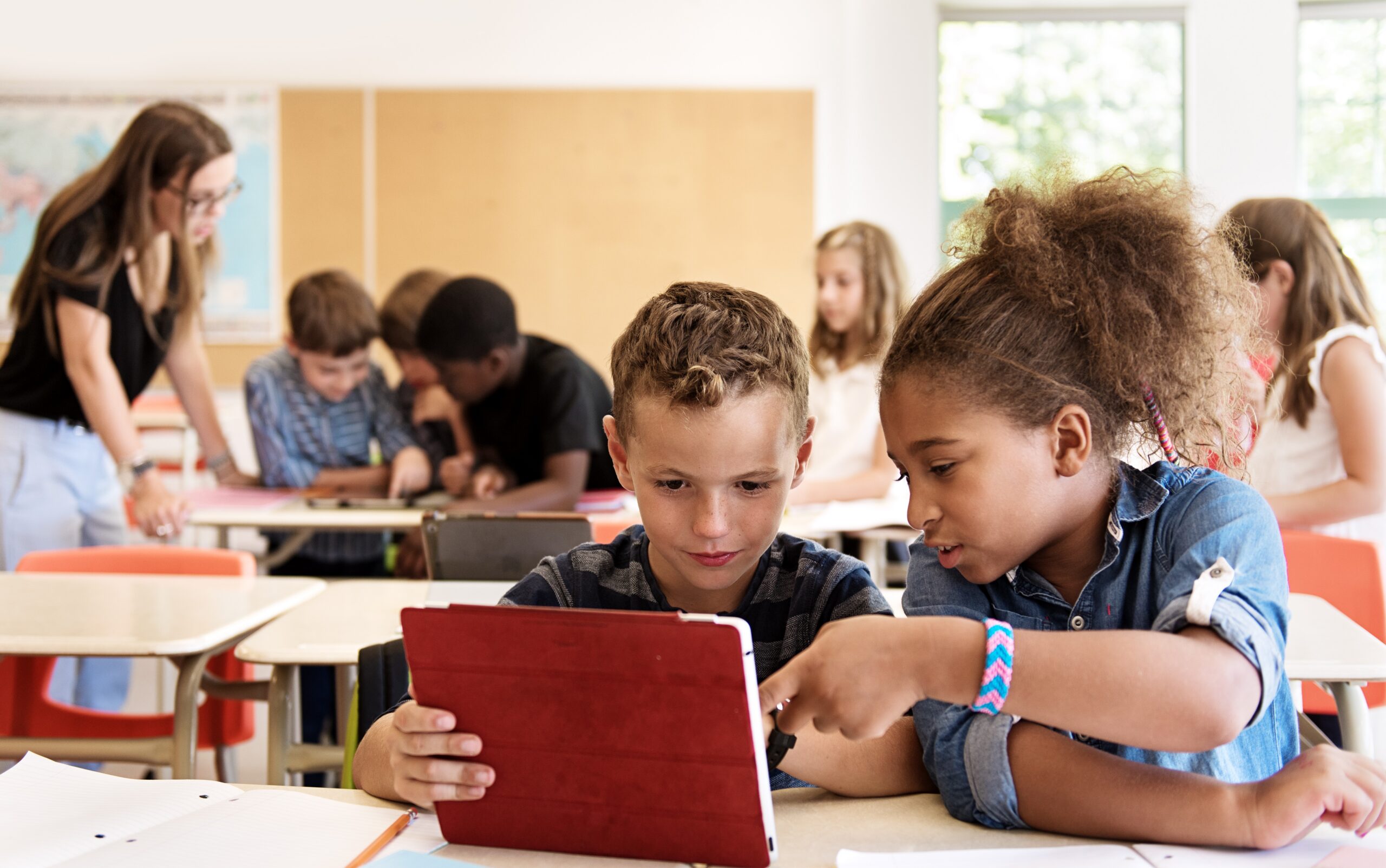 A boy wearing a striped t-shirt sits at a desk with a girl wearing a denim shirt reading a tablet together