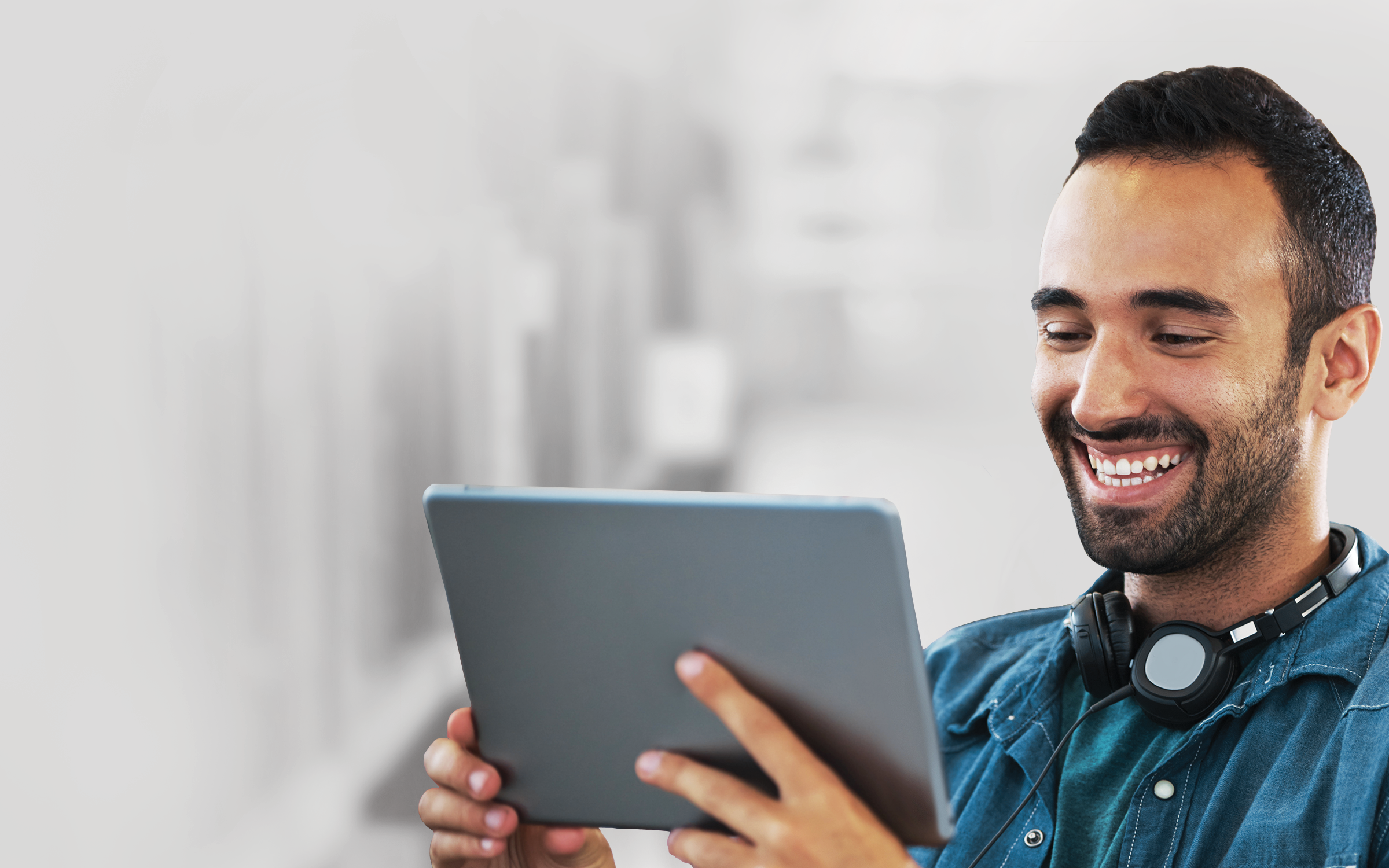 A man wearing a denim shirt smiles while he reads on a tablet wearing a pair of black wired headphones