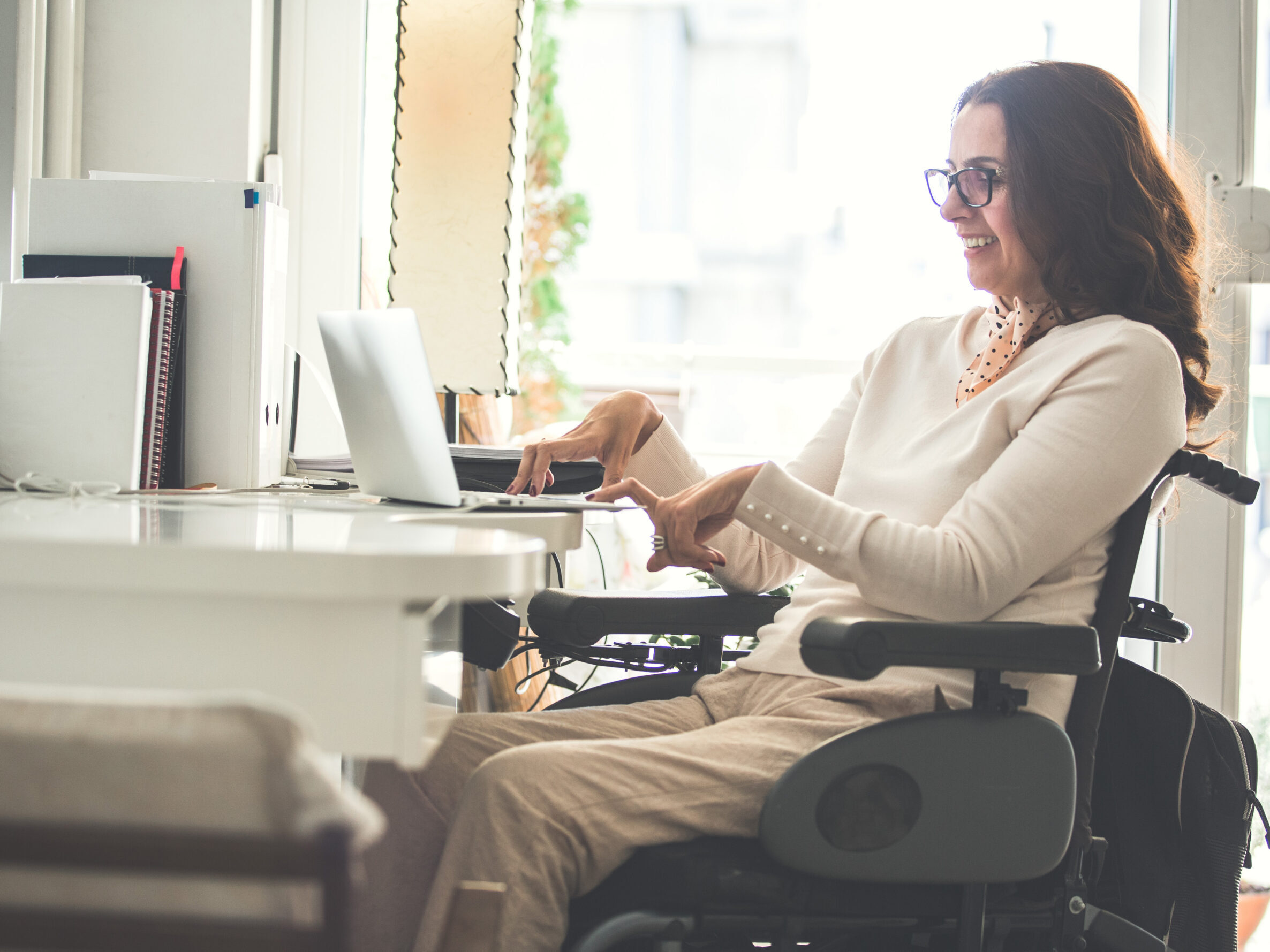 A woman wearing a white sweater and glasses sits in her wheelchair at her desk working on a laptop